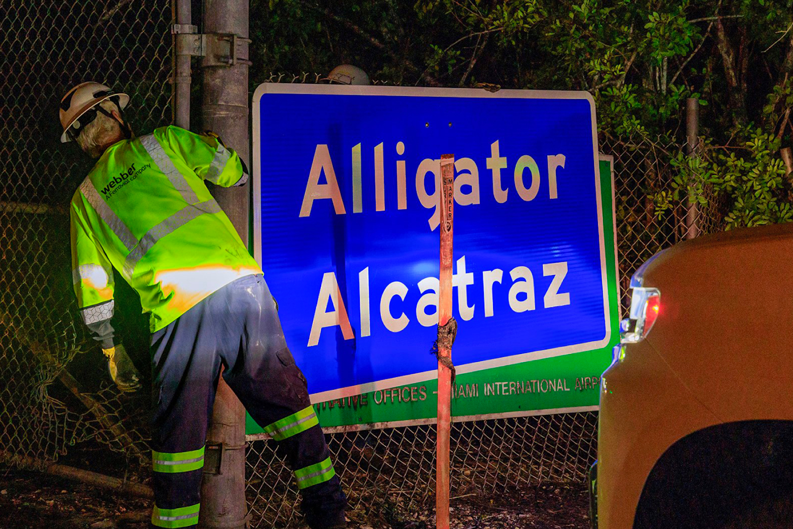 Man in a construction uniform leans against a blue sign that reads 'Aligator Alcatraz' at the entrance to Trump's Florida concentration camp.